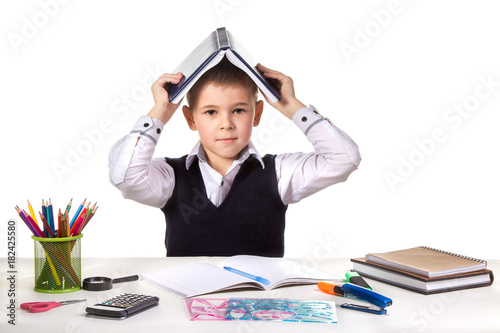 Smart serious excellent pupil at the desk with open book on the head on the white background