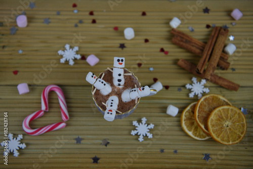 winter season food photography image with hot chocolate drink in a cup with mini marshmallows shaped as snowman with iced on smile and placed on kitchen table with cinnamon sticks and candy cane heart
