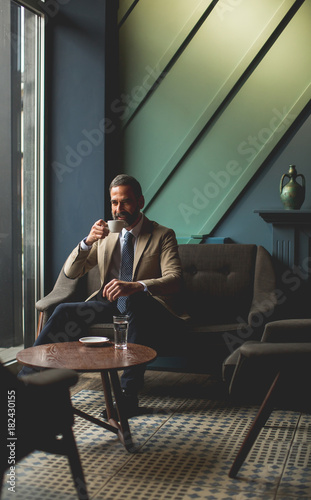 Handsome elegant serious businessman sitting in armchair and holding cup of espresso.