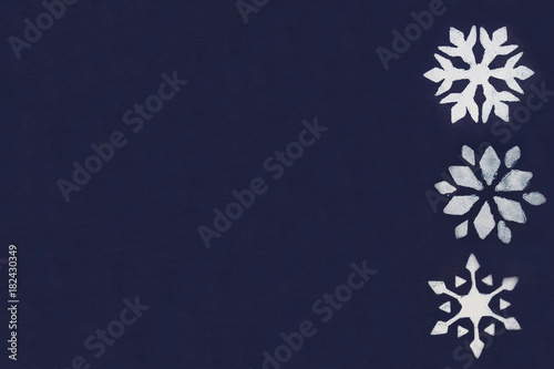 A few white snowflakes are painted through a stencil on a dark blue background. Copy space.