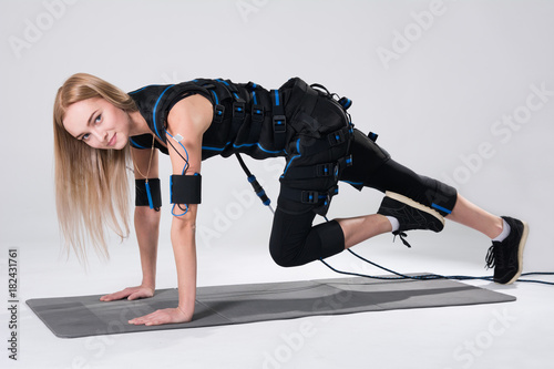 Beautiful blonde in an electric muscular suit for stimulation makes an exercise on the rug photo