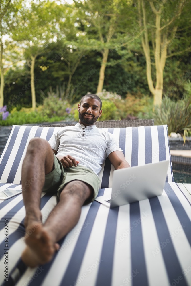 Fototapeta premium Smiling man using laptop while relaxing on lounge chair at porch