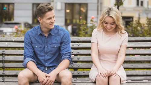 Shy blonde girl smiling, attractive guy flirting with beautiful woman on bench photo