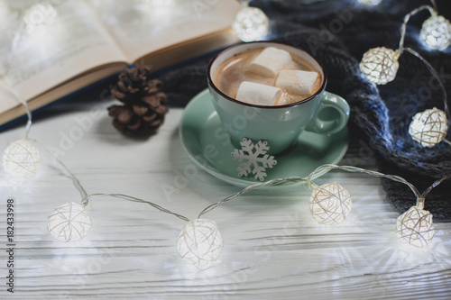 Cozy winter home. Cup of cocoa with marshmallows, warm knitted sweater, open book, Christmas garland on a white wooden table. Atmosphere of a pleasant evening for reading.