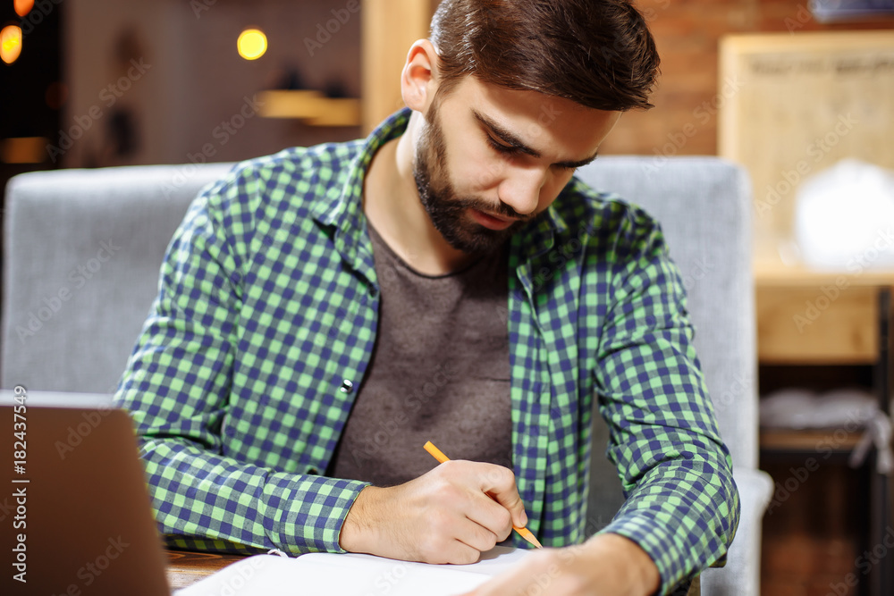 Confident young manager sitting at cafe table and writing down creative ideas in notepad, blurred background Confident young manager sitting at cafe table and writing down creative ideas in notepad, blurred background