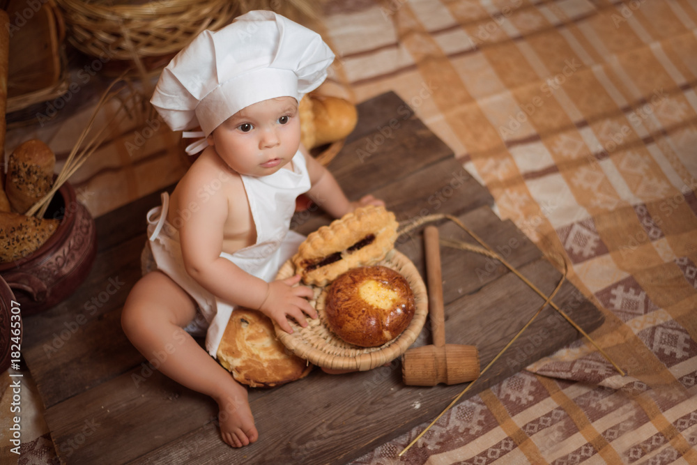 Happy little child in the cook costume at the kitchen sitting on the table among baskets with tasty rolls, buns and bread. Copy space Happy little child in the cook costume at the kitchen sitting on the table among baskets with tasty rolls, buns and bread. Copy space