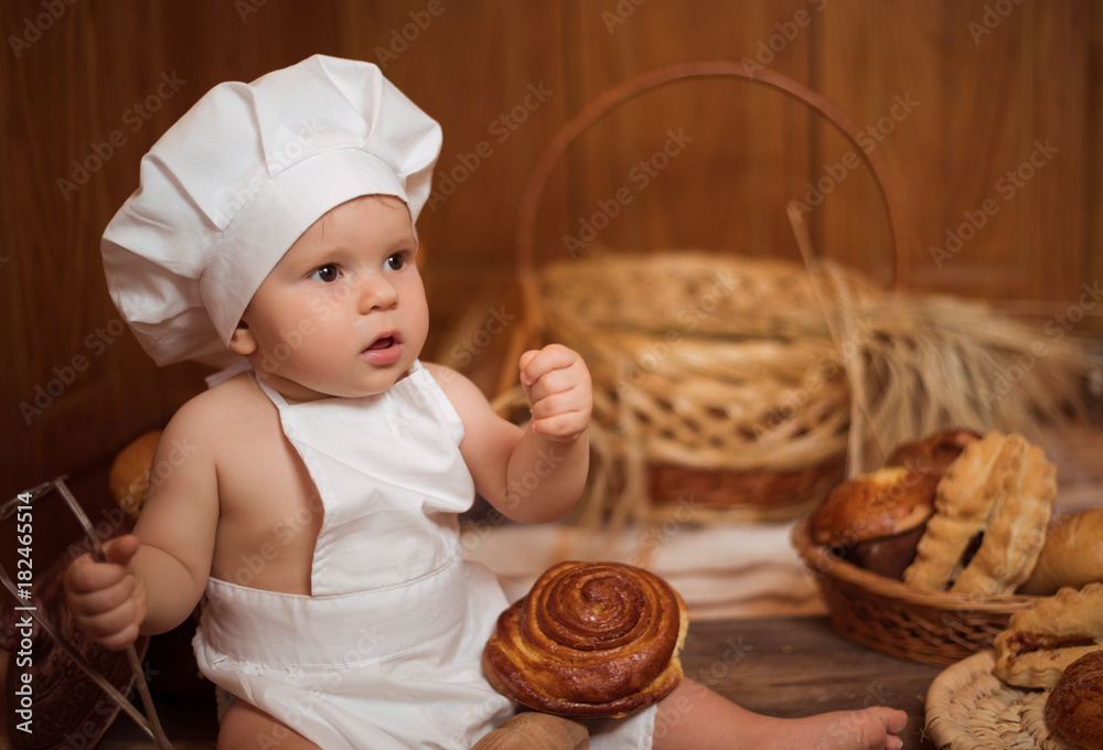 Happy little child in the cook costume at the kitchen sitting on the table among baskets with tasty rolls, buns and bread. Copy space Happy little child in the cook costume at the kitchen sitting on the table among baskets with tasty rolls, buns and bread. Copy space