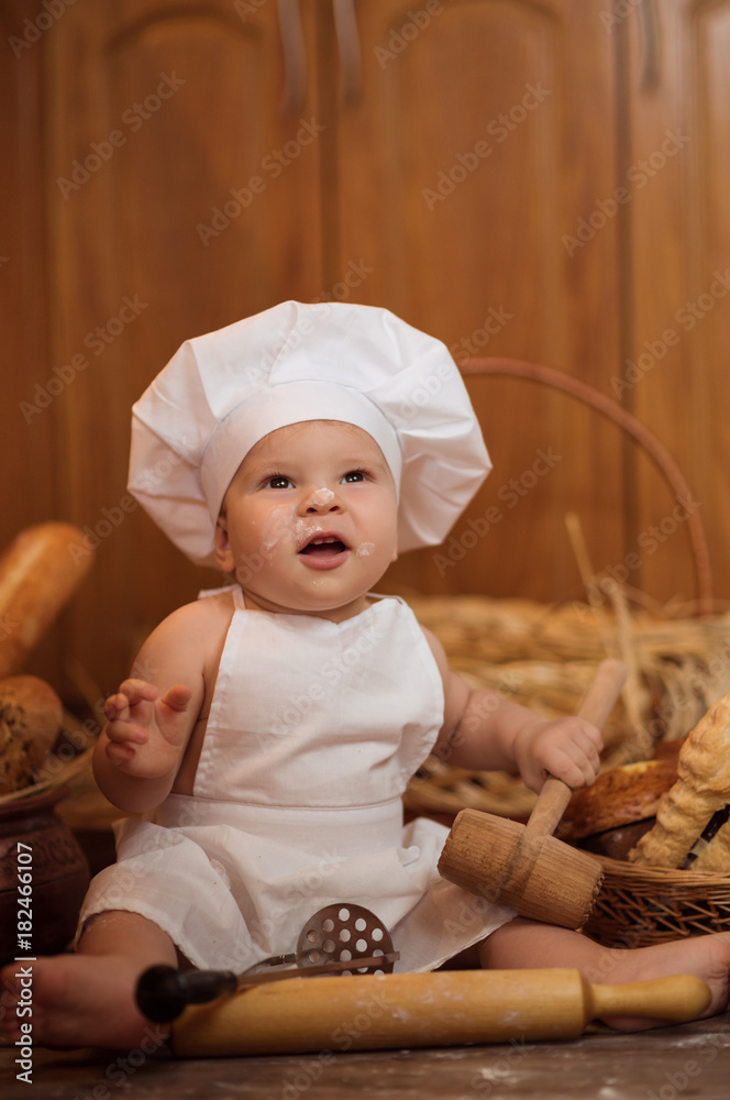little boy in a cook costume among a baking bread rolls of flour confectionery on a brown wooden floor sits on a wooden background little boy in a cook costume among a baking bread rolls of flour confectionery on a brown wooden floor sits on a wooden background