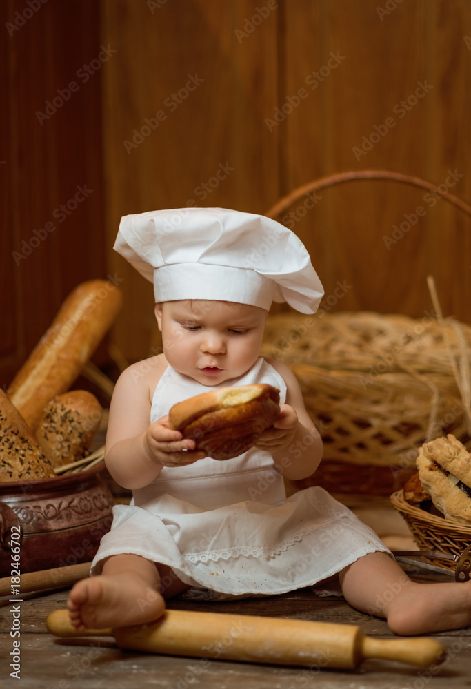 Small child in hat as a cook eating a croissant in the background of baskets with rolls and bread. Copy space Small child in hat as a cook eating a croissant in the background of baskets with rolls and bread. Copy space