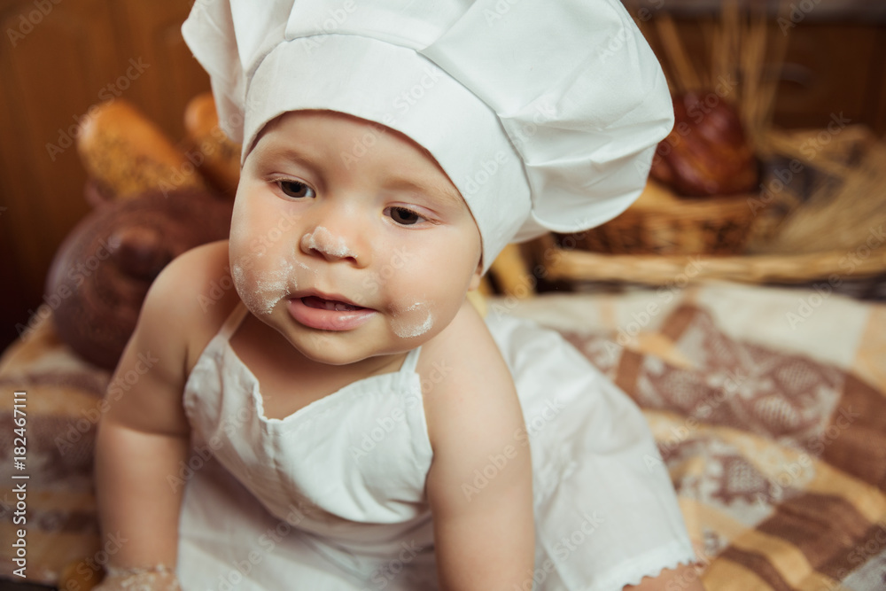 Happy little child in the cook costume at the kitchen sitting on the table among baskets with tasty rolls, buns and bread. Copy space Happy little child in the cook costume at the kitchen sitting on the table among baskets with tasty rolls, buns and bread. Copy space