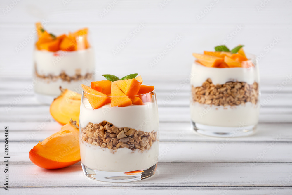 Persimmon creamy trifle in beautiful glasses, fresh ripe fruit slices on white wooden background. Healthy vegetarian food. Delicious dessert. Close up photography. Selective focus. Persimmon creamy trifle in beautiful glasses, fresh ripe fruit slices on white wooden background. Healthy vegetarian food. Delicious dessert. Close up photography. Selective focus.