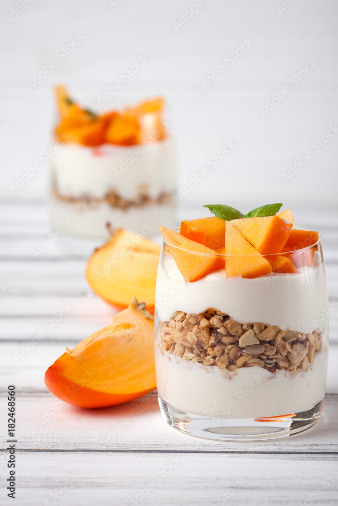 Persimmon creamy trifle in beautiful glasses, fresh ripe fruit slices on white wooden background. Healthy vegetarian food. Delicious dessert. Close up photography. Selective focus. Persimmon creamy trifle in beautiful glasses, fresh ripe fruit slices on white wooden background. Healthy vegetarian food. Delicious dessert. Close up photography. Selective focus.