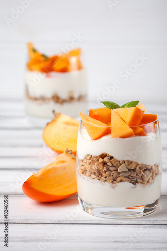 Persimmon creamy trifle in beautiful glasses, fresh ripe fruit slices on white wooden background. Healthy vegetarian food. Delicious dessert. Close up photography. Selective focus.