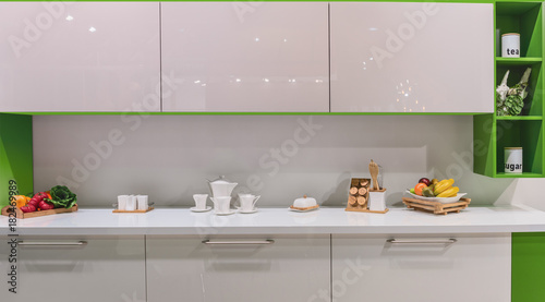 Fragment of the kitchen in a modern style with dishes  fruits and vegetables on a white table top. Glaringly glossy facades and bright green furniture details.