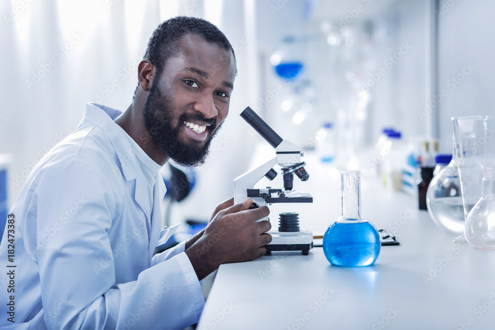 Positive mood. Cheerful nice handsome scientist sitting in front of the microscope and looking at you while enjoying his job Positive mood. Cheerful nice handsome scientist sitting in front of the microscope and looking at you while enjoying his job