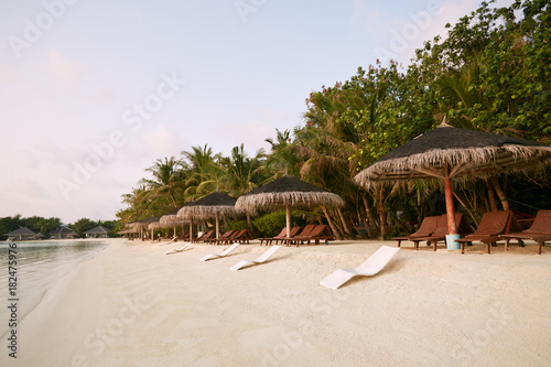 Beach chairs under straw umbrellas. Indian ocean coastline on Maldives island. White sandy beach and calm sea. Travel and vacation concept.