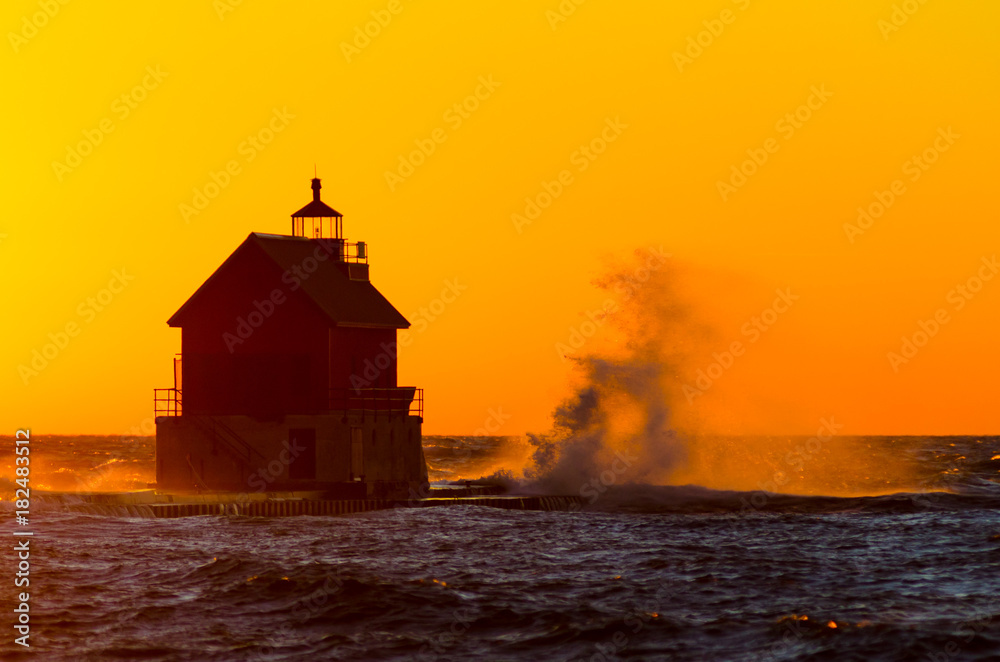 A bright yellow sunset highlights waves splashing against the lighthouse and pier at Grand Haven, Michigan