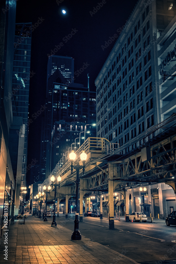 City street at night in Chicago's Loop with elevated train station, illuminated buildings, cars and moon..