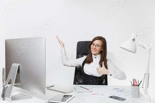 Smiling business woman sitting at the desk, working at computer with documents in light office, showing thumb up, pointing her hand at white background with copy space for your advertisement or text