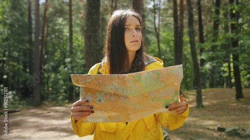 Young beautiful worried woman in yellow coat and backpack looking for the route with a help of a map in the middle of the forest. Got lost. Outside