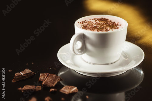 Closeup image of white cup full of latte coffee at dark background.