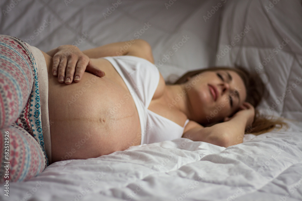 In bed, a pregnant young girl sleeps and has her hand on her stomach. Focus on the abdomen In bed, a pregnant young girl sleeps and has her hand on her stomach. Focus on the abdomen