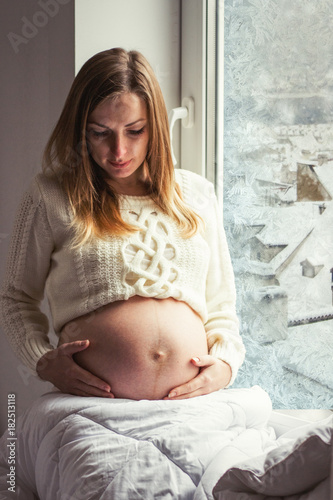 Pregnant young girl holds her hands in her belly. Outside the window is winter