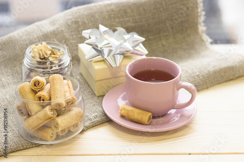 A festive still life with a pink tea cup with a saucer, a clear bowl of fresh cookies, a light gift box with a shiny silver bow, dry flowers in a jar on a linen cloth, a light wooden background