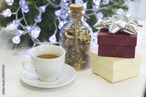 A festive bright morning with a white porcelian cup of hot black tea, a glass clear bottle of dry flowers, a light gift box and a purple gift box with a silver bow, Christmas tree with white fairy