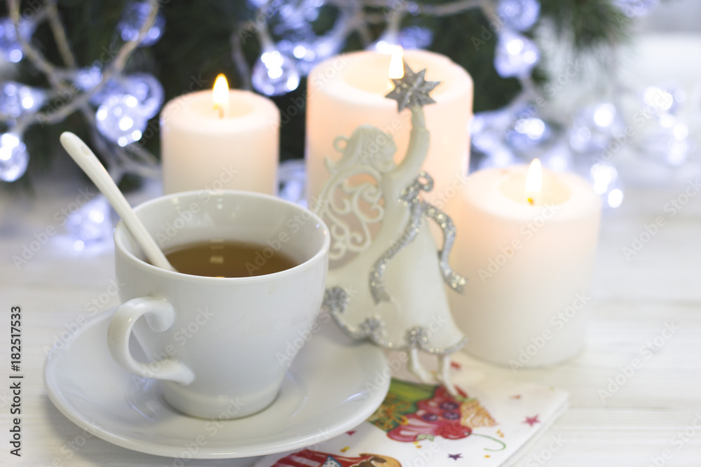 A festive still life with a white tea cup, a spoon and a saucer, a white porcelian angel, tree white burning candles, a blurred Christmas tree with fairy lights on the back, light background