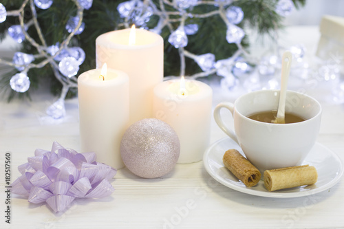 A festive still life with a white tea cup, a spoon and a saucer, a shiny pink Christmas-tree ball, a purple bow, tree white burning candles, a blurred Christmas tree with fairy lights on the back