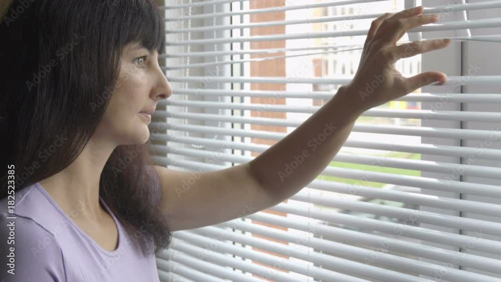 Young beautiful curious woman looks through the window through blinds. The girl is watching out the window. The brunette woman.