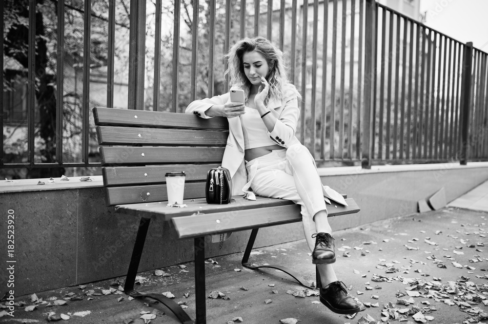 Obraz premium Stylish curly blonde model girl wear on white with cup of coffee and mobile phone on hands posing at bench at fall.
