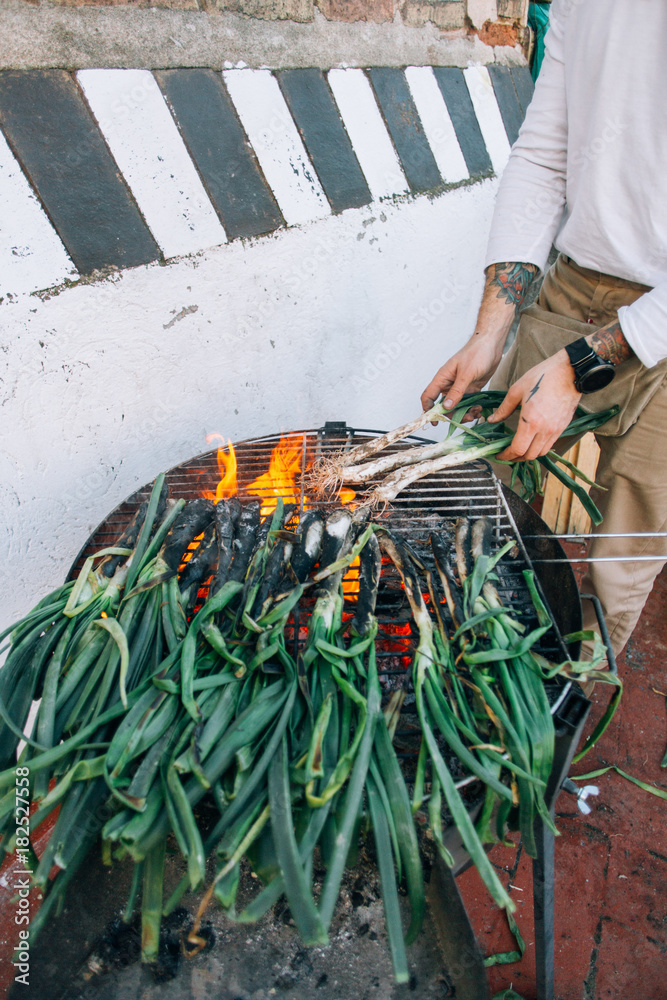 Obraz premium Man in white tshirt and with authentic arm tattoos, makes barbecue on grill on outside terrace of spring onions, calcots or cambray as traditional spanish, catalan or mexican food