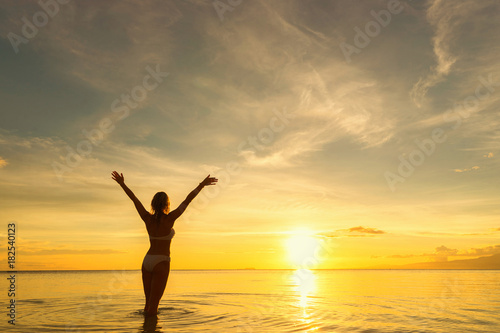 Back of young woman standing in water with raised arms and looking at sea and sunset.