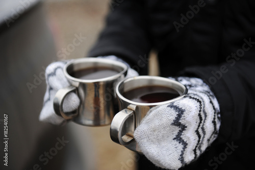 Two metal cups with coffee in hands in mittens close-up on car background. Brewing coffee in the open air