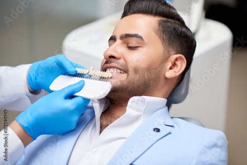 Shot of a handsome young man on a medical appointment at the dentist office professional dentist holding dentures comparing color and shade of teeth medicine healthcare whitening. 