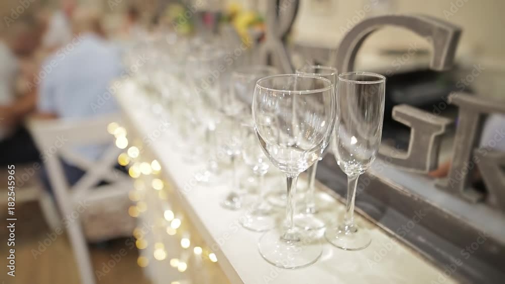 A lot of empty glasses on white table cloth. Yellow lights of a garland and unrecognizable people are sitting at the table on a blurred background.