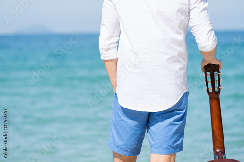 Back view of man holding guitar and standing on the beach with blue sea and blue sky bacjground. Travel relax vacation concept.