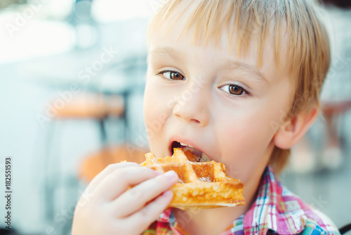 Cute little boy eats tasty belgian waffle on summer terrace.