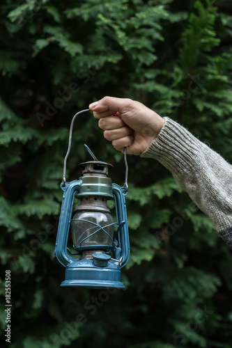 Old lantern held in a hand in front of a dark green tree © Anna