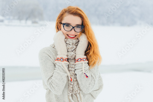 Beautiful smiling red head woman with glasses is wrapped in the scarf during the snowfall in the forest. Half-length horizontal portrait.