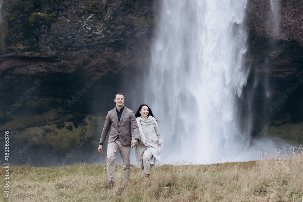 Bride and groom walking on the grass feald in Iceland. Couple hold hands and smiling. They are look so happy. Woman wear beautiful linen dress and knitted scarf. Icelandic waterfall on the background.