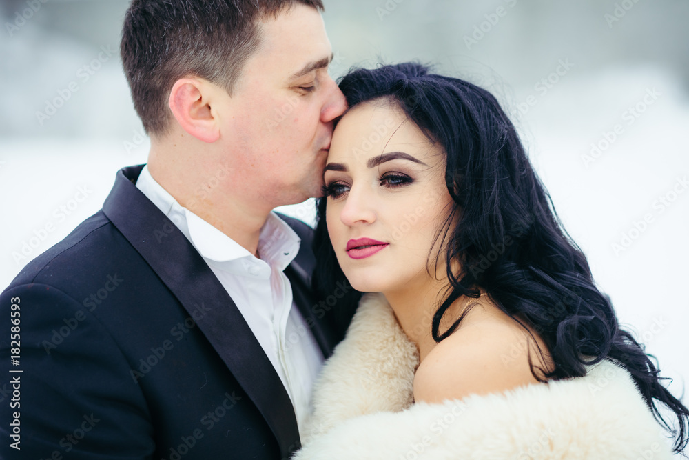 Horizontal portrait of the groom kissing the beautiful serious brunette bride into forehead. Winter time. Horizontal portrait of the groom kissing the beautiful serious brunette bride into forehead. Winter time.
