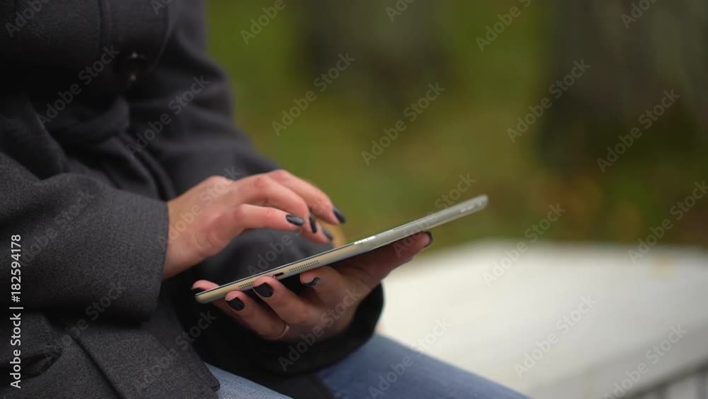 Young woman using tablet computer while sitting on bench in autumn park. Close-up of female hands touching of tablet computer. Student with a Tablet Sitting on the Bench and Using Laptop in a Autumn