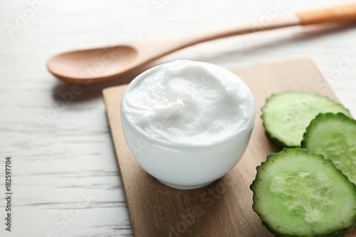 Bowl of body cream with sliced cucumber on wooden board