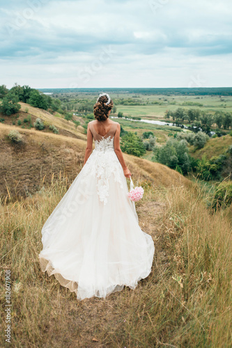 A young girl, the bride in a long wedding dress, is turned her back and stares into the distance to the river and a beautiful landscape.