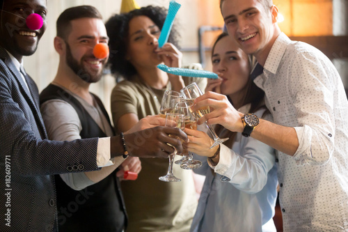 Multiracial young people in party hats clinking glasses celebrating New years day eve or birthday, diverse friends looking at camera having fun holding champagne, blowing whistles at celebration
