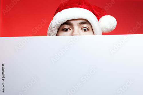Christmas selling, new year discounts and poster. Man in santa claus hat looking on blank white place for text. Guy with brown dark eyes on red white background © tavrox