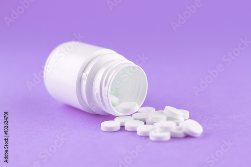 Close up white pill bottle with spilled out pills on purple background with copy space. Focus on foreground, soft bokeh. Pharmacy drugstore concept